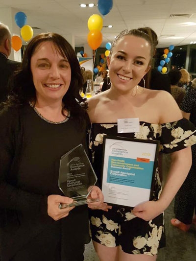 Two women smile at the camera. One is holding a trophy and the other is holding a certificate
