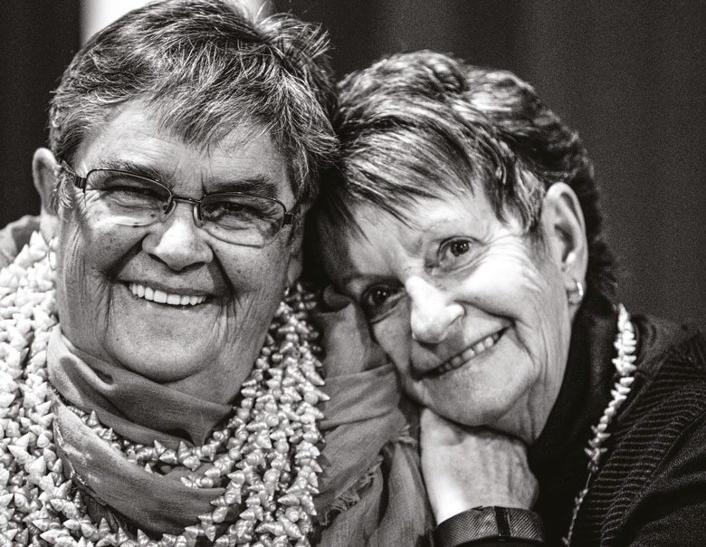 Two women lean against each other, smiling at the camera
